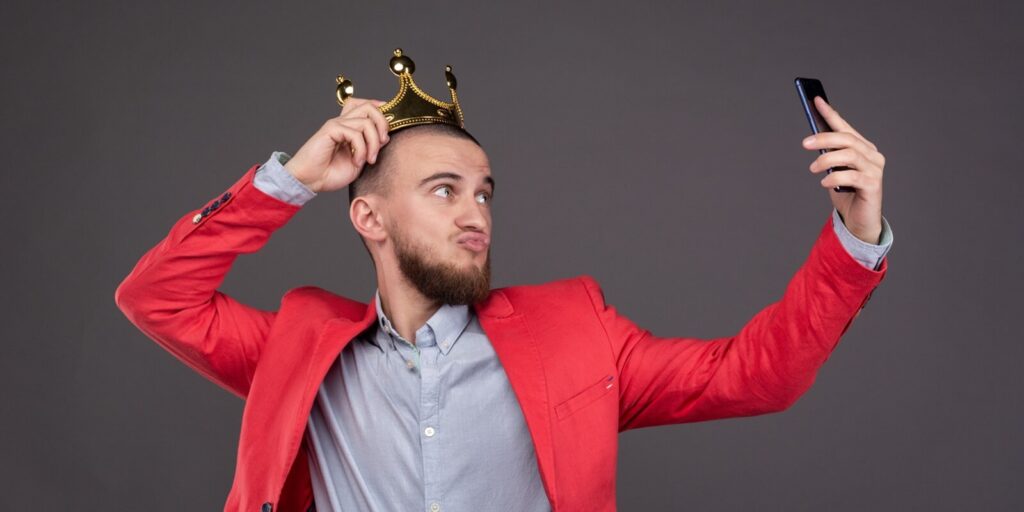 young bearded handsome man in gold crown taking selfie looking at smartphone
