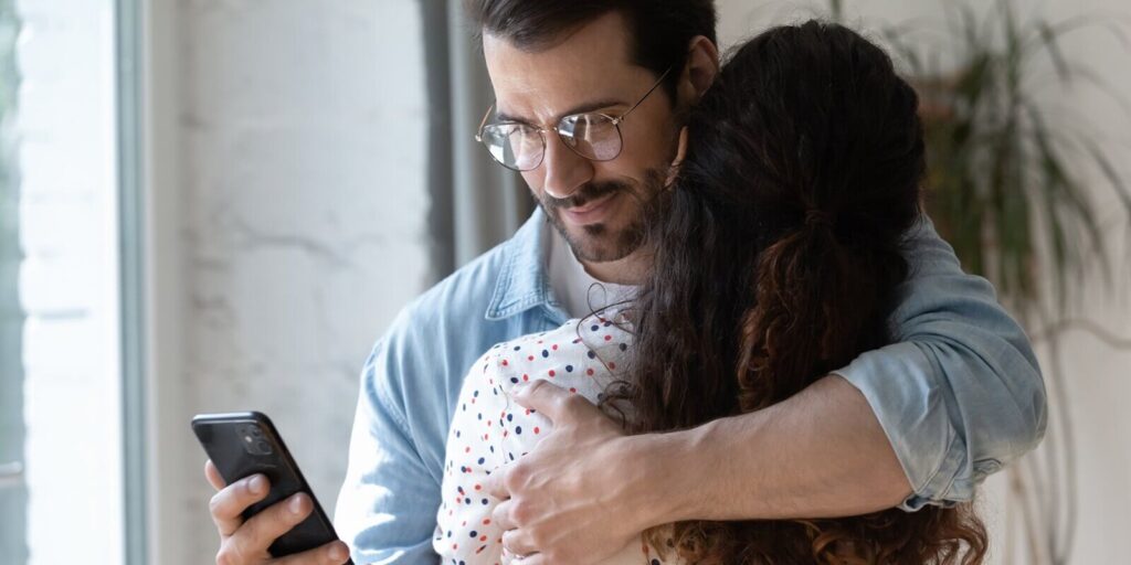 dishonest young man cuddling loving woman, using smartphone behind back